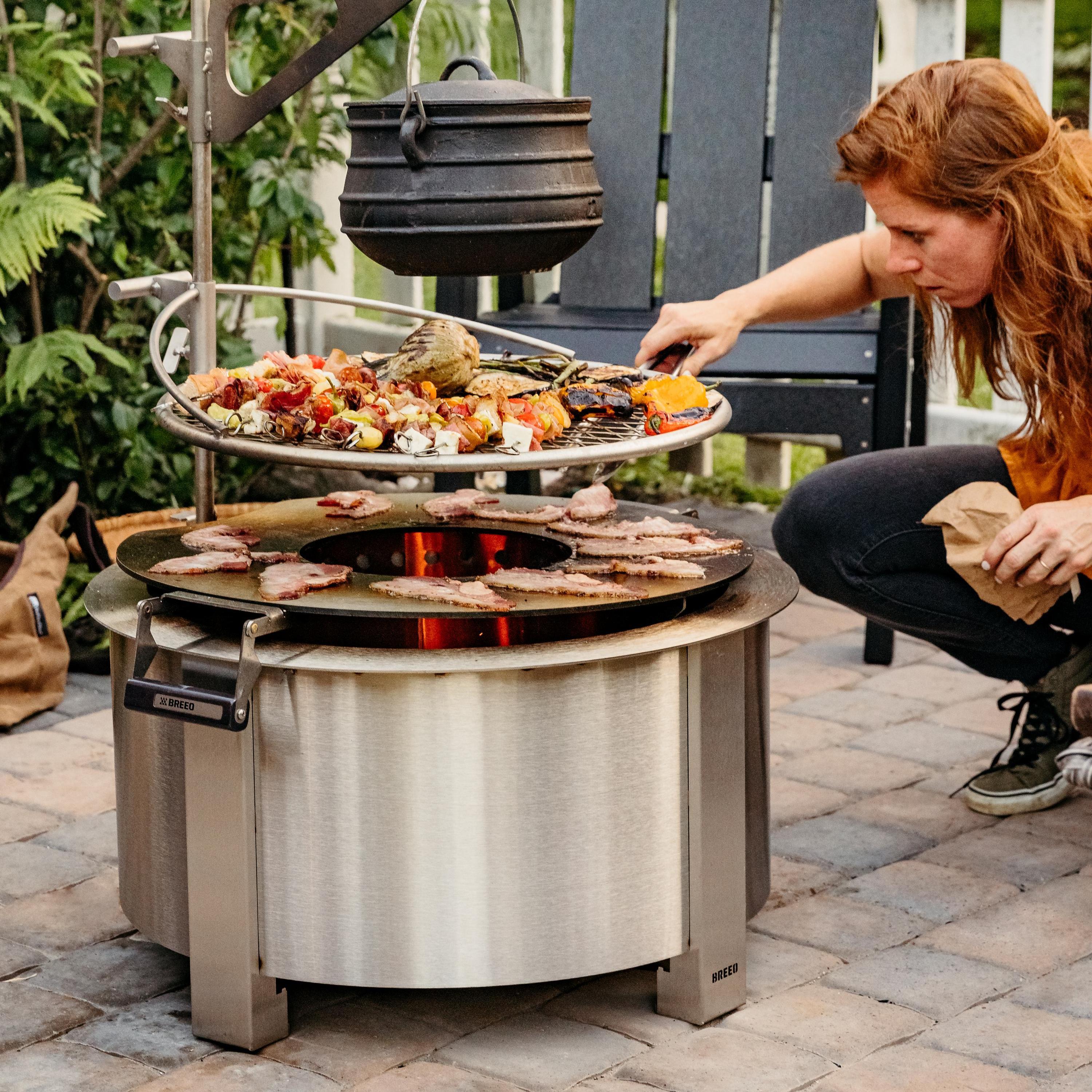 Person cooking food over tiered breeo firepit on patio with breeo chair