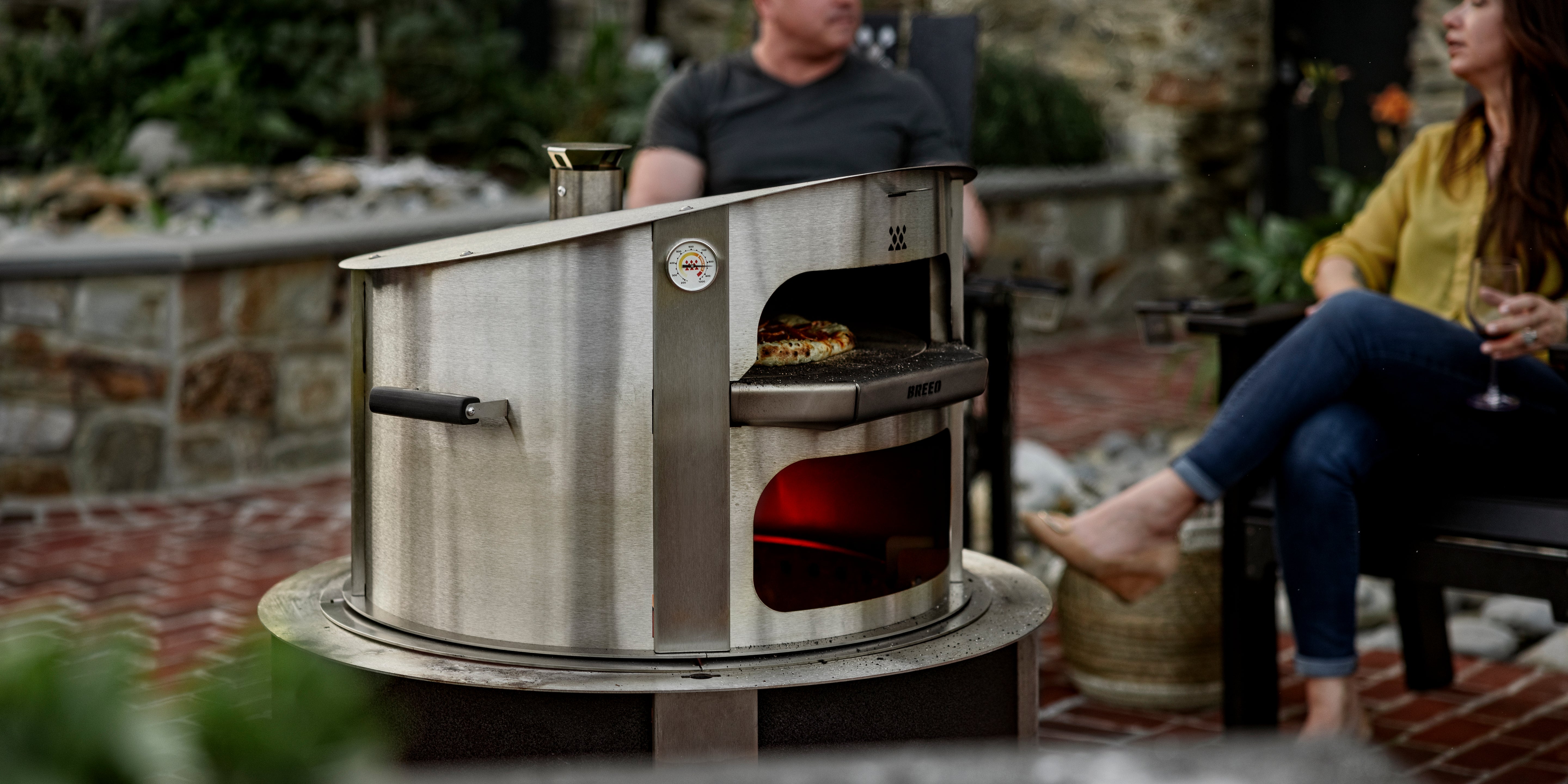 People sitting in breeo x series chairs around a breeo pizza oven with pizza cooking