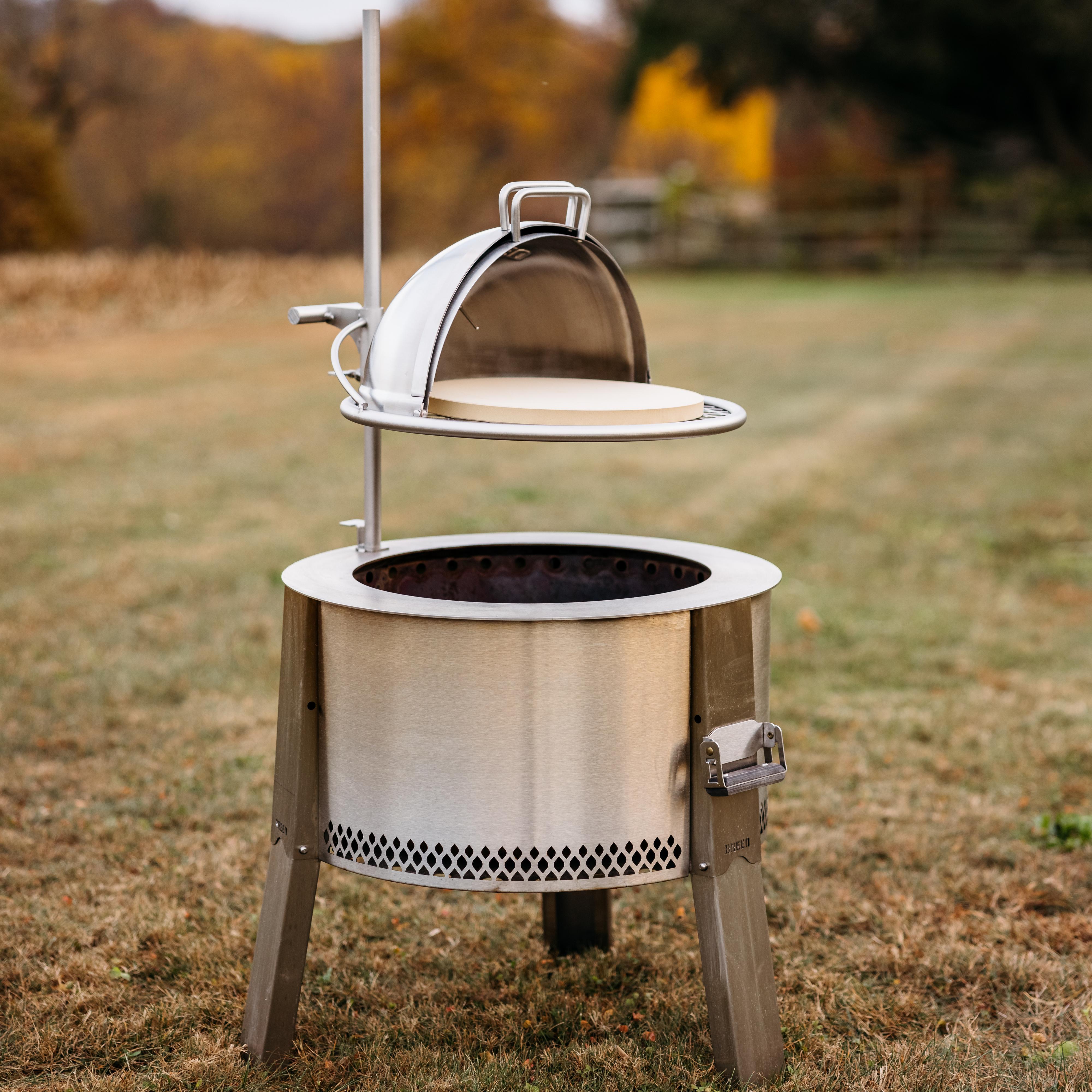 Breeo stainless steel fire pit with adjustable cooking system, featuring a dome lid lifted above a ceramic pizza stone, set on a grassy outdoor field in autumn