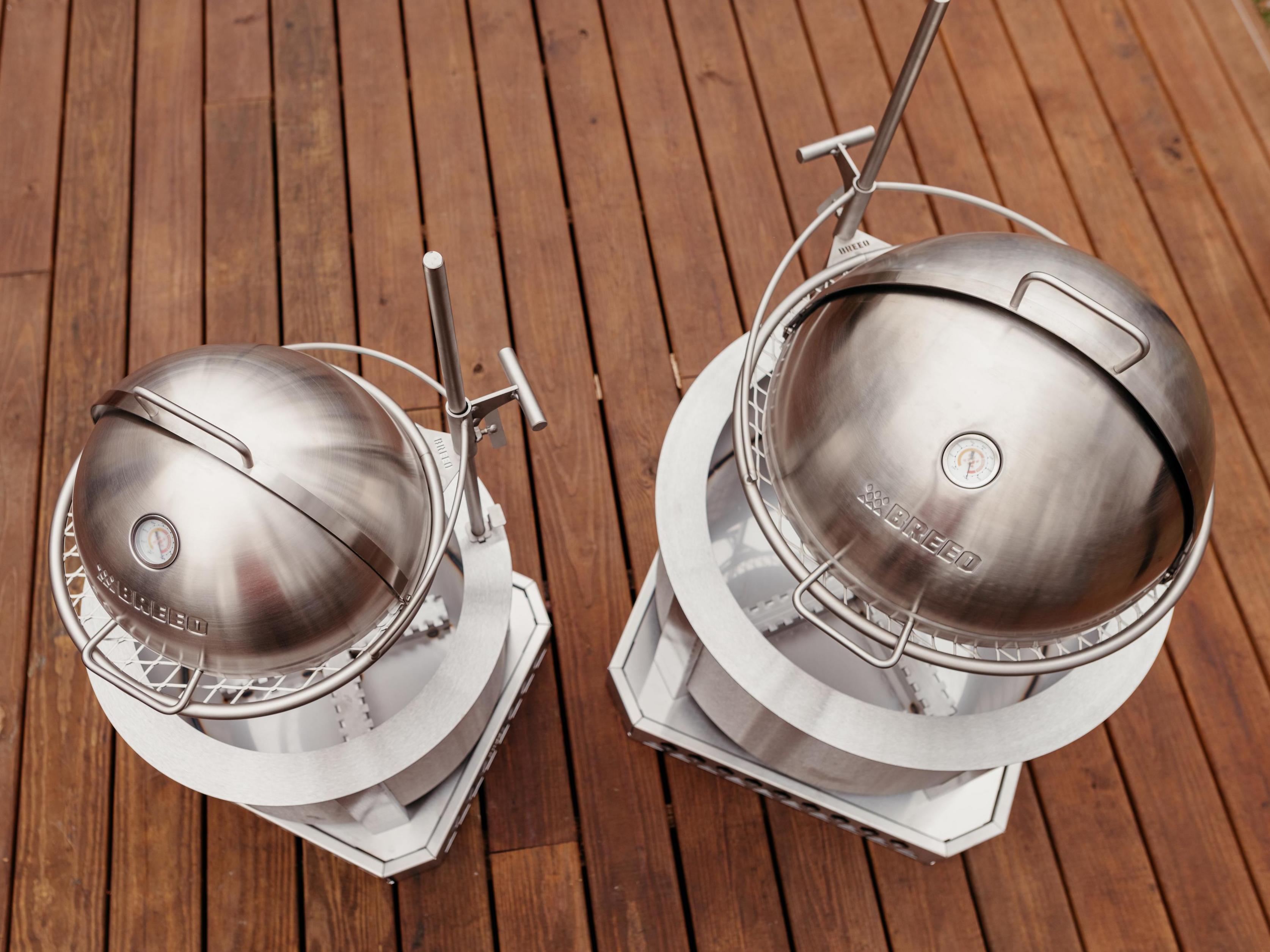 Top-down view of two Breeo live-fire grills with stainless steel dome lids and Outpost grill systems on a wooden deck, showcasing size comparison.