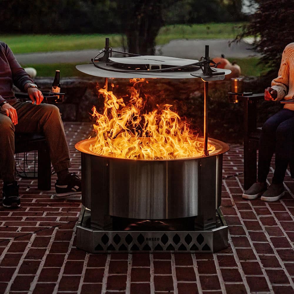 Two users sit around an X 30 on a Breeo base sitting on a brick patio with a roaring fire and the Heat Deflector installed.