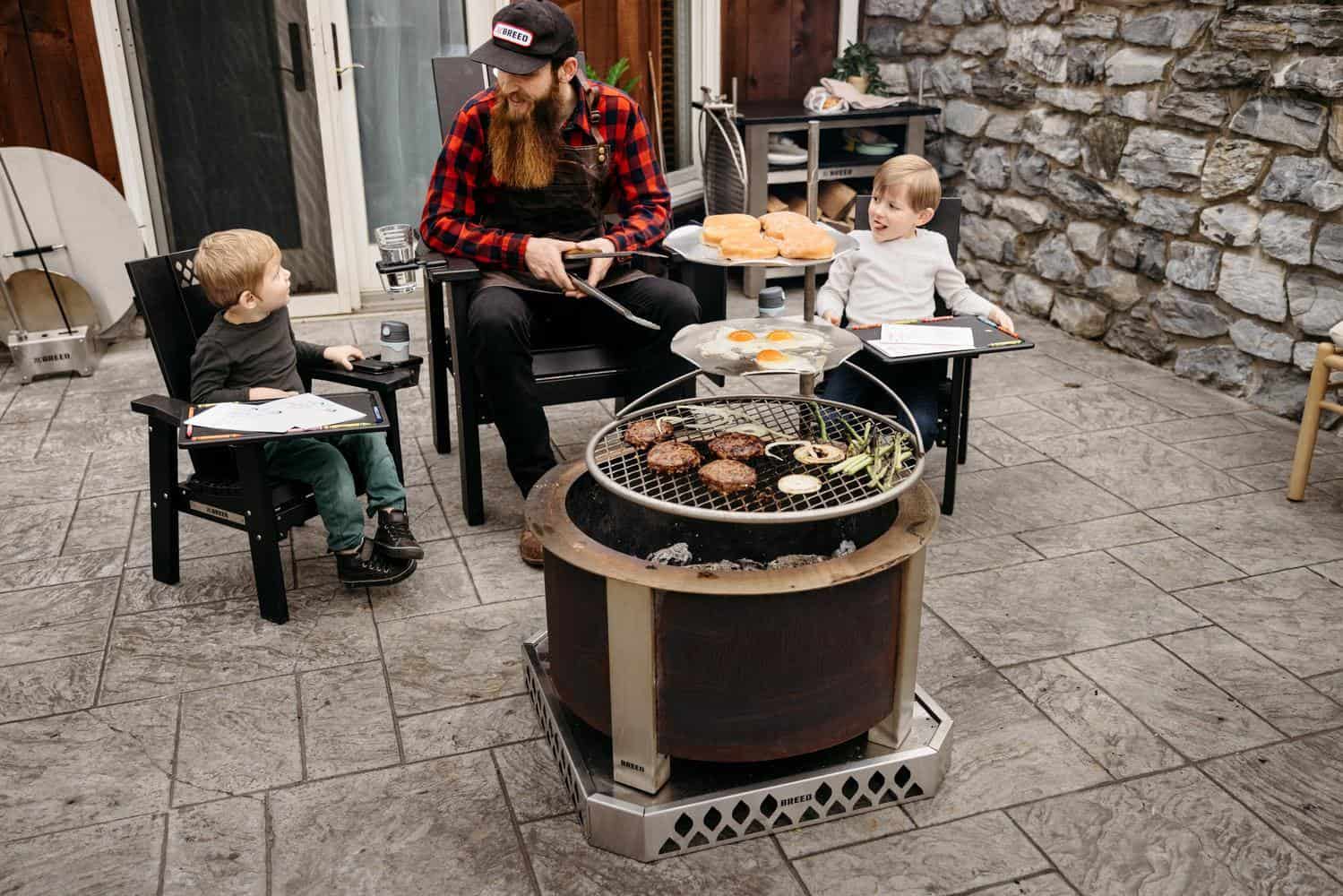 In this image a Dad is sitting around Breeo Firepit with his sons while grilling using an Outpost Grill and Outpost Sear Discs.