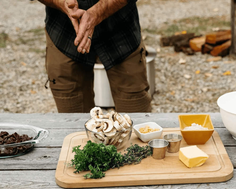 An image of a man standing behind fresh ingredients, preparing to cook his Venison Liver and Mushroom Gravy. 