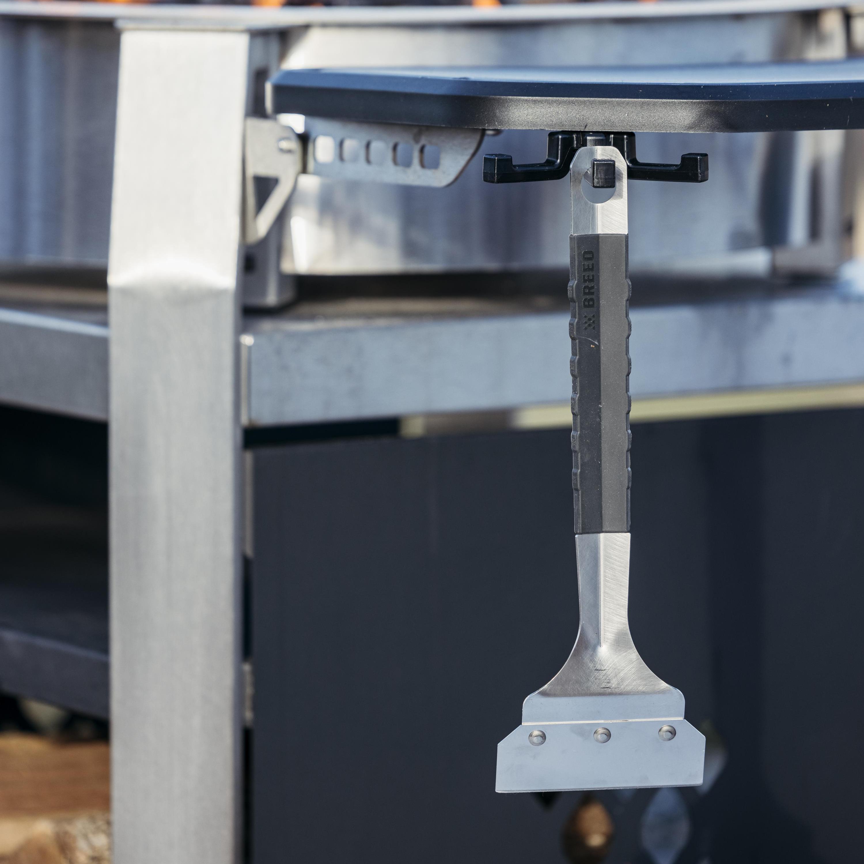 A close-up of a breeo stainless steel griddle scraper attached to a griddle, with a metal handle.