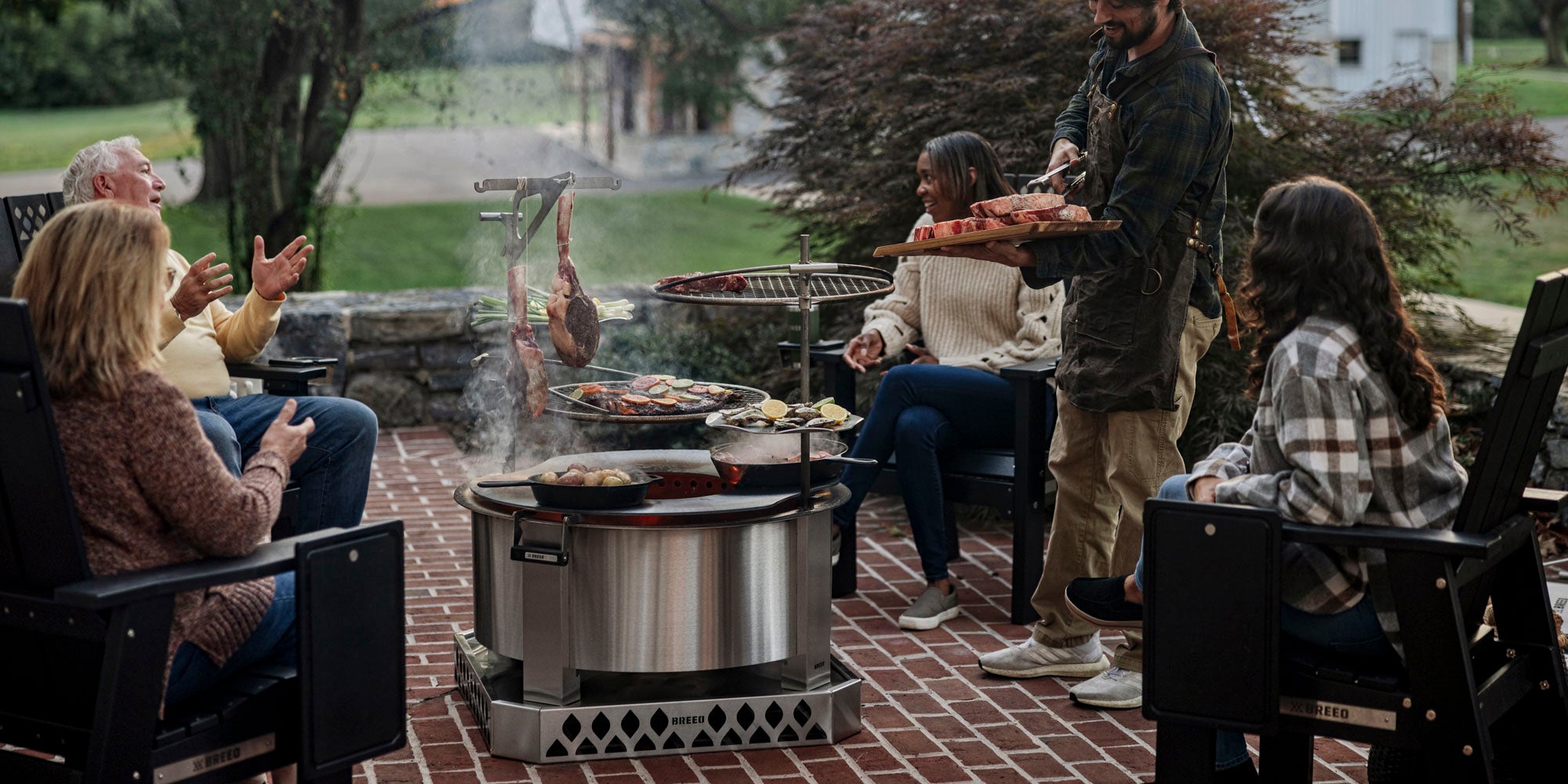 People gathered around stainless breeo smokeless fireit cooking meat and other food outdoors on brick and stone patio