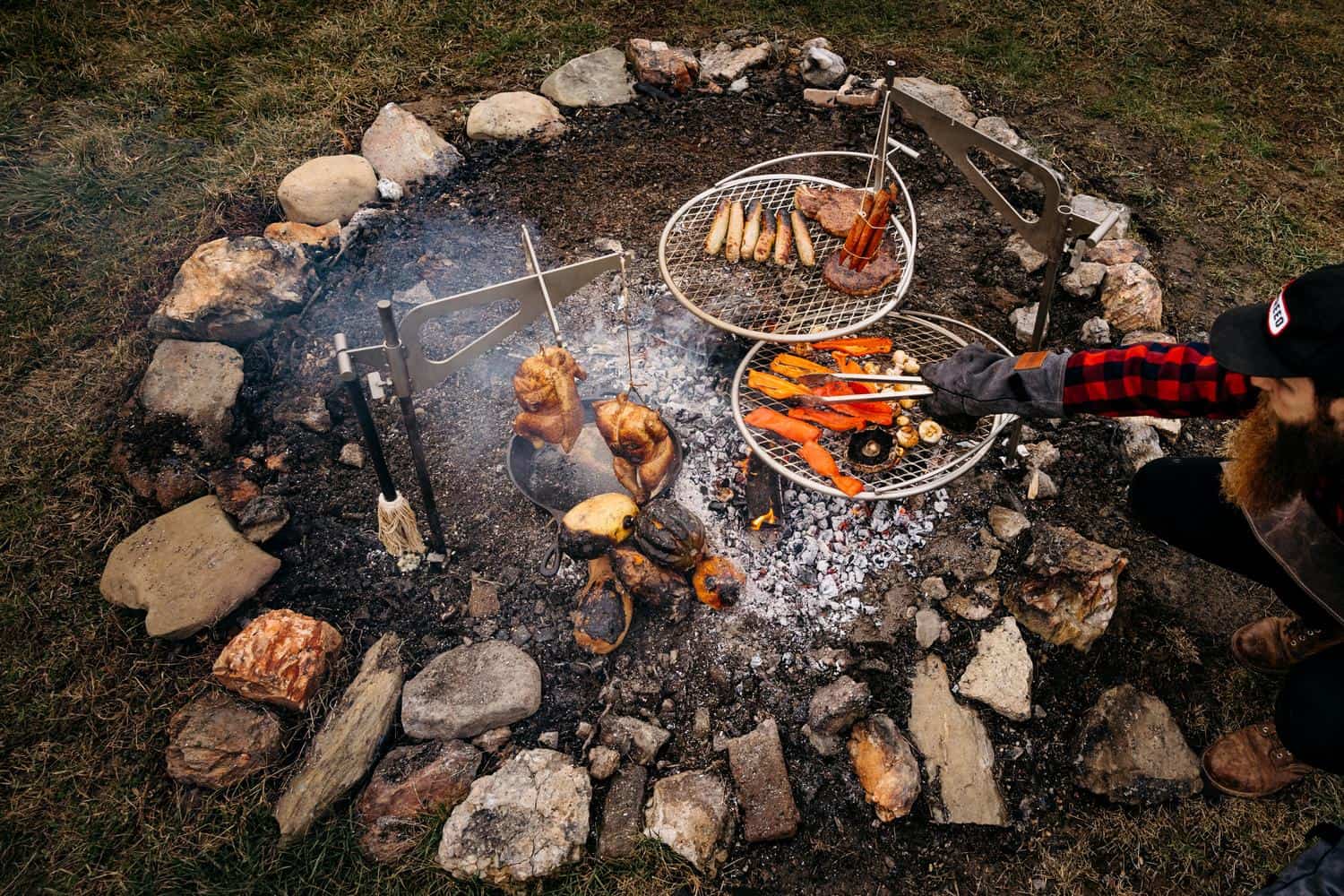 An image of a man grilling on two Outpost Grills and an Outpost Kettle Hook with Rotisserie chickens hanging from it. 